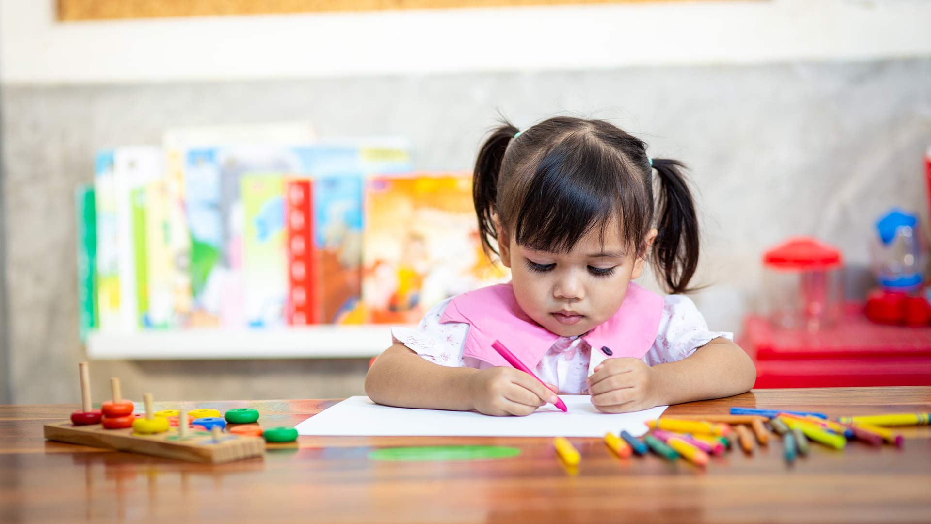 Junges Mädchen malt mit einem rosa Wachsmalstift im Kindergarten