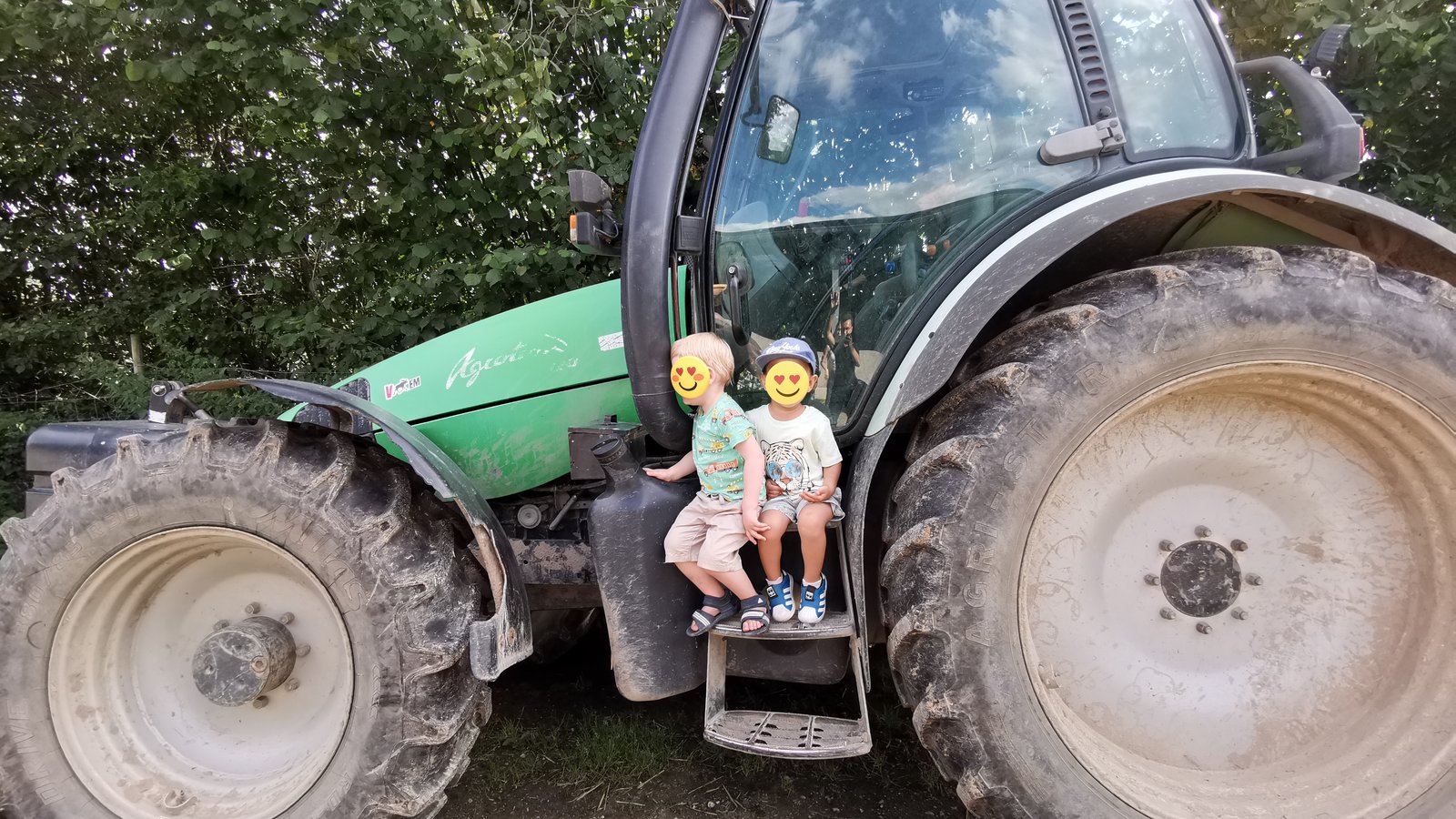 Two young children sitting on the step of a big green tractor next to a huge rear wheel